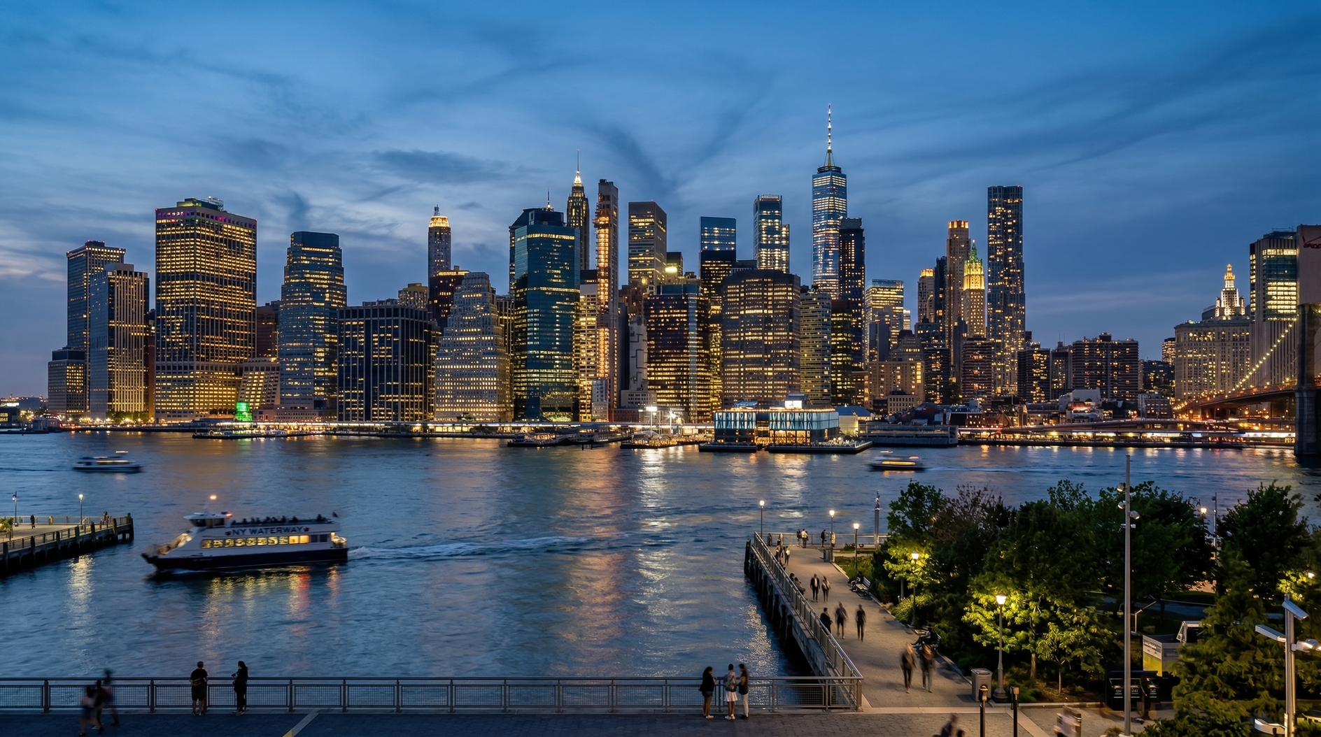 Illuminated Manhattan skyline at dusk viewed from Brooklyn waterfront with reflections on East River